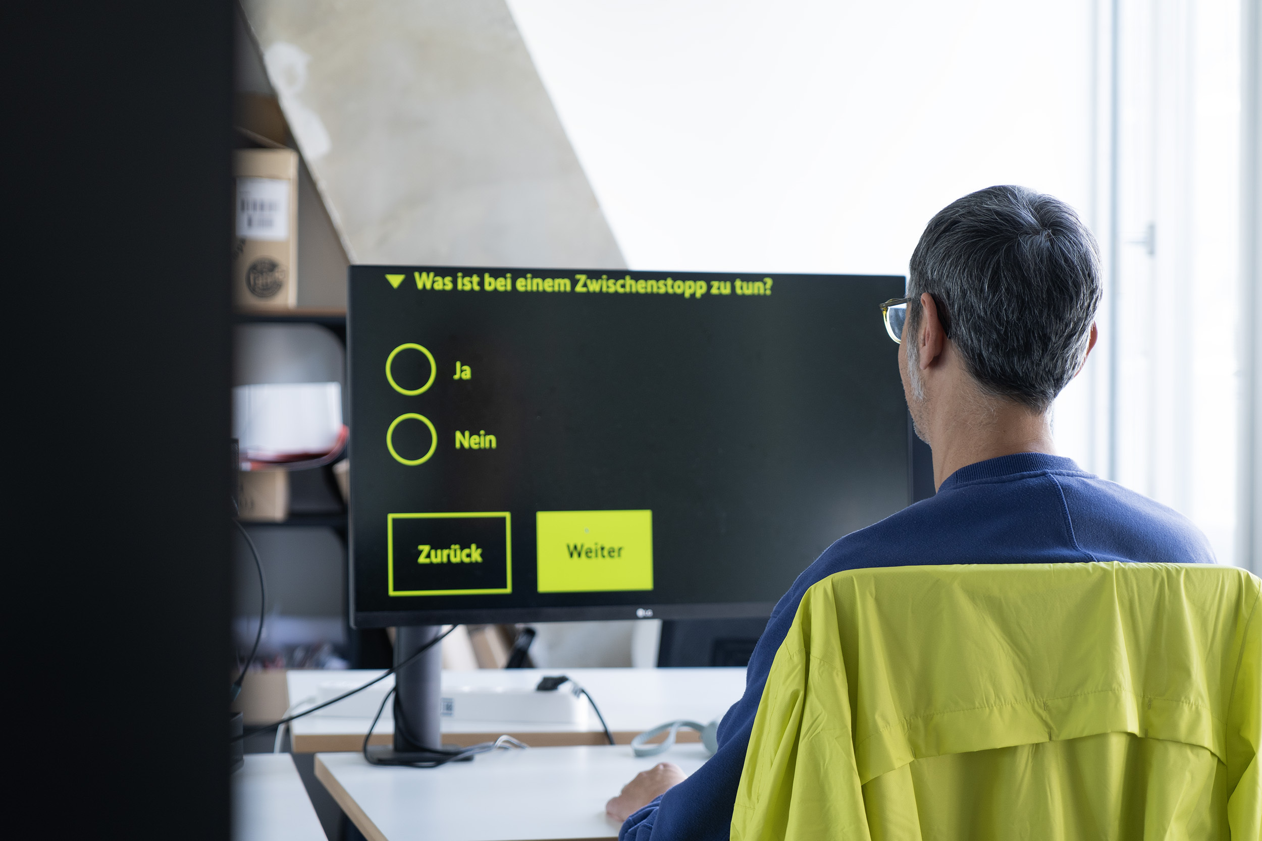 Ein Mann sitzt vor einem Monitor, auf dem stark vergrößert eine Multiple-Choice-Frage auf Deutsch angezeigt wird: „Was ist bei einem Zwischenstopp zu tun?“ mit den Optionen „Ja“ und „Nein“ sowie Buttons „Zurück“ und „Weiter“.