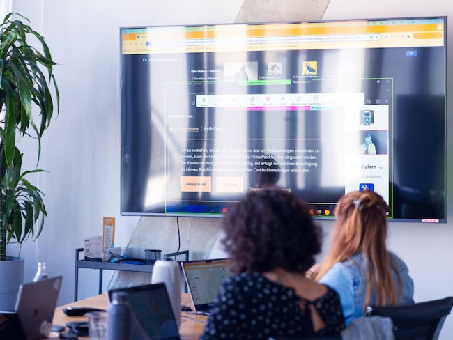 Zwei Frauen sitzen vor Laptops in einem modernen Büro, während ein großer Bildschirm eine Videokonferenz und eine Webseite anzeigt. Eine Zimmerpflanze steht links im Raum.