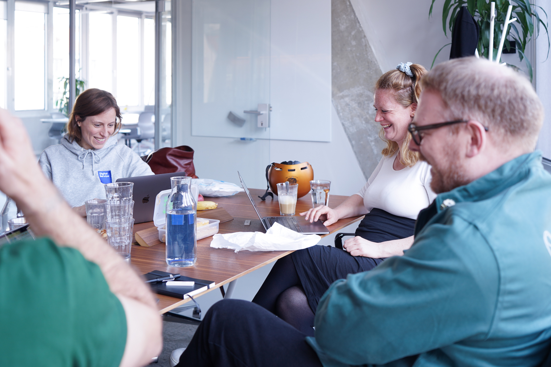 Vier Personen sitzen an einem Tisch in einem Meetingraum. Auf dem Tisch stehen aufgeklappte Laptops und Getränke. Drei der vier Personen sind erkennbar am lachen.