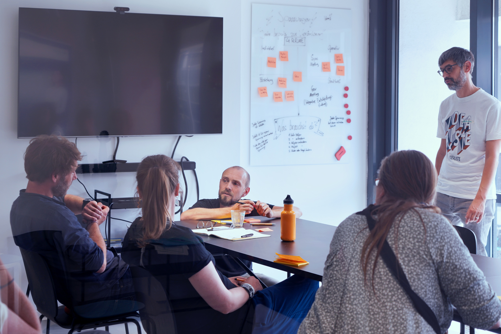 Fünf Menschen in einem Meetingraum. Eine Person steht, eine kniet an den Tisch gelehnt. Die anderen drei sitzen. Alle sind einer Person zugewandt, die gerade etwas sagt.