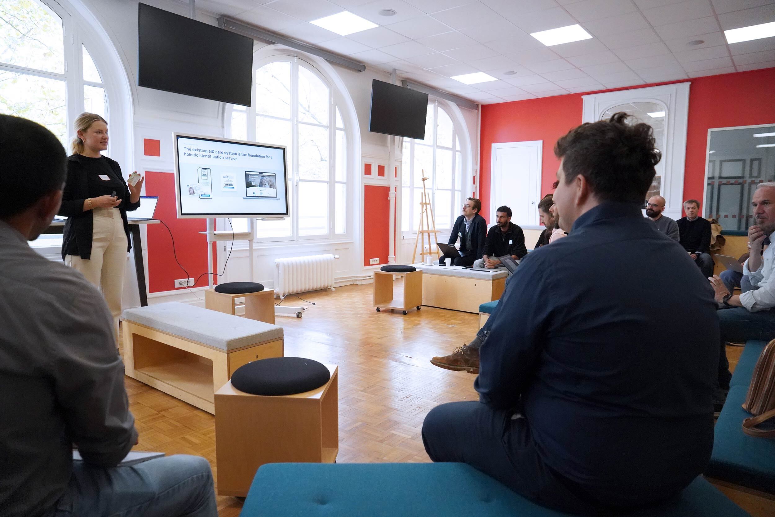 Eine Gruppe von Menschen in einem modernisierten Raum mit farbigen Wänden – sie alle sitzen auf Holzbänken und blicken auf eine blonde Frau, die neben einem Bildschirm steht und zu ihnen spricht