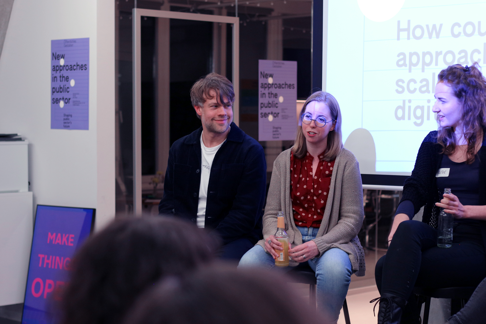 Drei Menschen, zwei Frauen und ein Mann, mit helleren Hauttönen sitzen vor einer Leinwand und Zuhörenden und schauen einander an – um sie herum hängen Poster auf denen in englischer Sprache steht: „Neue Ansätze im öffentlichen Sektor“ – und „Tue Dinge im Offenen“