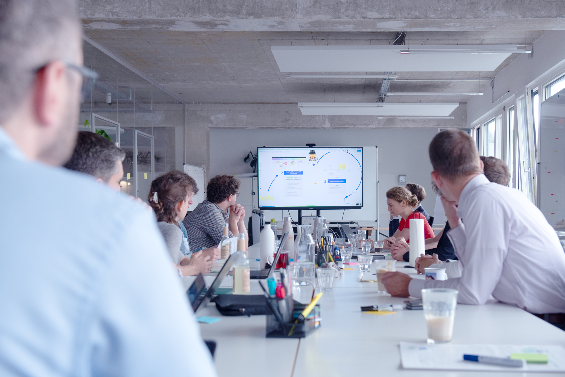 Eine größere Gruppe sitzt um einen Konferenztisch und diskutiert, am vorderen Ende ist auf einem Bildschirm eine Präsentation zur schrittweisen Weiterentwicklung des Digitalcheck zu sehen.