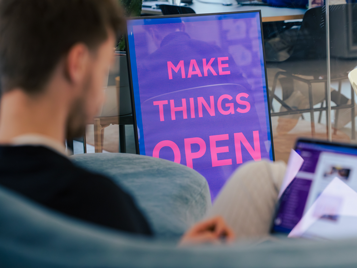 Eine Person sitzt auf einem Sofa und schaut auf den Bildschirm eines Laptops auf ihrem Schoß. Im Hintergrund lehnt ein Bild gegen eine Glaswand. Auf dem Bild steht „MAKE THINGS OPEN“.