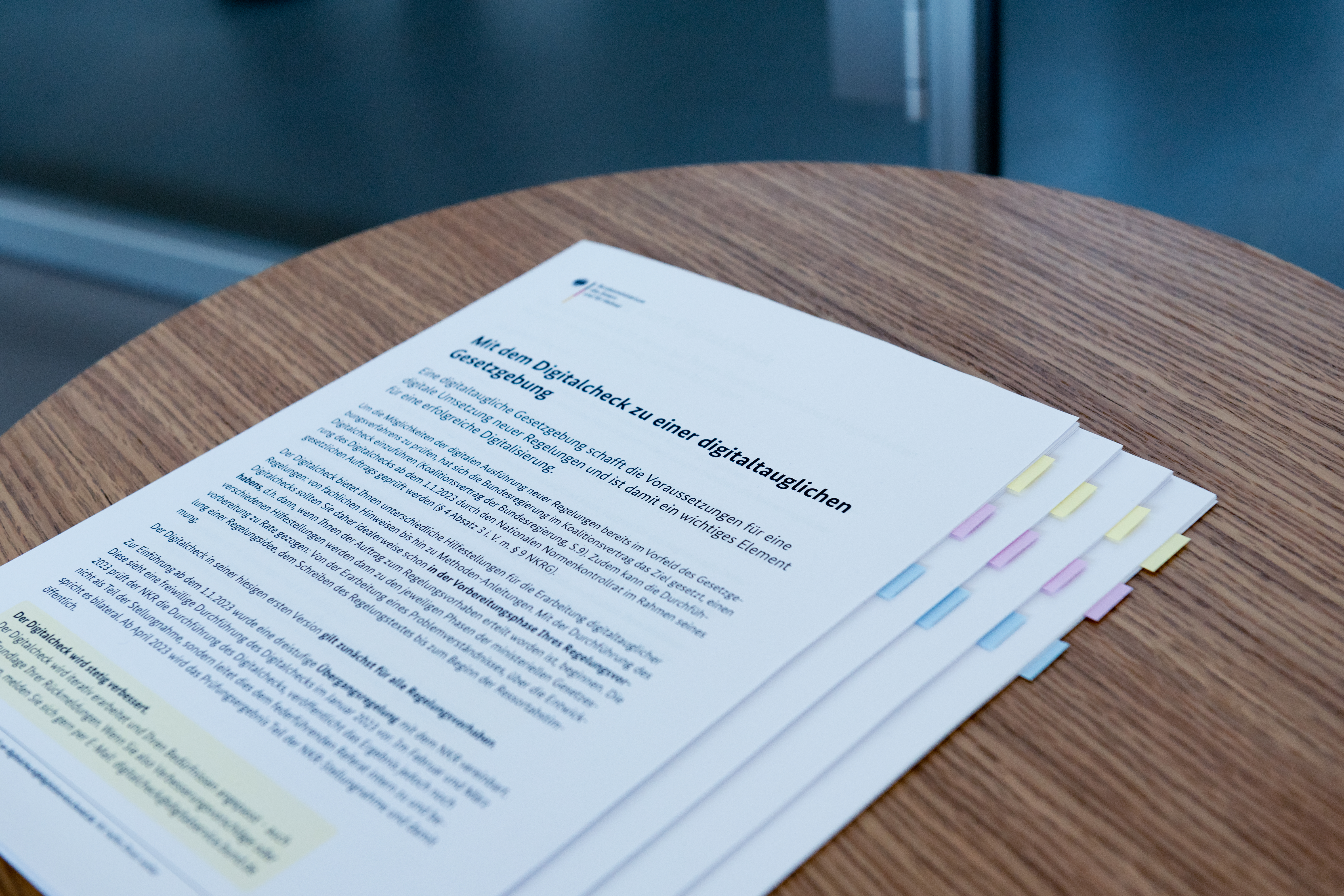 Ein Stapel bedruckter Dokumente mit dem Titel „Mit dem Digitalcheck zu einer digitaltauglichen Gesetzgebung“ liegt auf einem runden Holztisch; die Seiten sind mit farbigen Registermarken versehen.