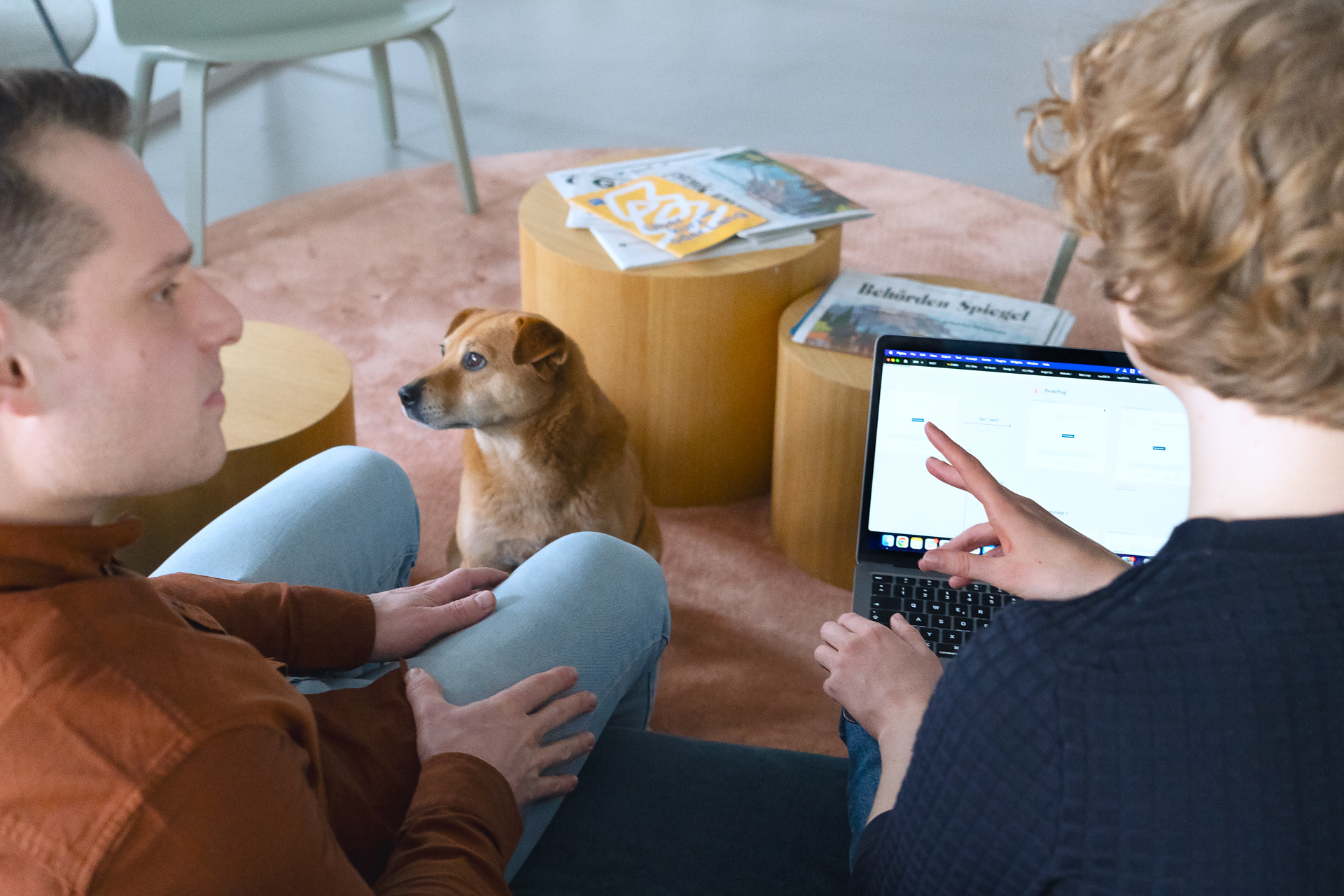 Zwei Personen unterhalten sich auf einer Couch sitzend. Eine Person hat einen Laptop auf dem Schoß. Zwischen den beiden auf dem Boden sitzt ein Hund.