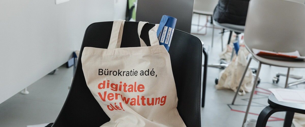 Ein Beutel liegt auf einem Stuhl. Auf ihm steht „Bürokratie adé, digitale Verwaltung olé!“