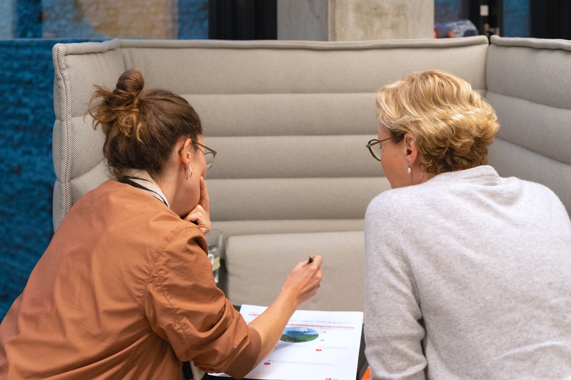 Projektpartnerin Sabine und Fellow Anne beugen sich über ein Dokument. Ihre Schultern und Hinterköpfe sind zu sehen. Sie sitzen in einer abgeschirmten Sitzecke.