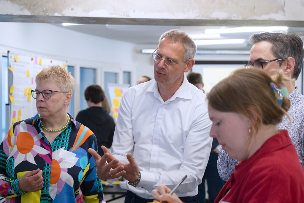 Vier Personen besprechen sich, eine davon gestikuliert mit den Händen, eine notiert sich etwas. Im Hintergrund stehen weitere Personen vor Flipcharts mit Textausdrucken, die mit Post-its beklebt sind.