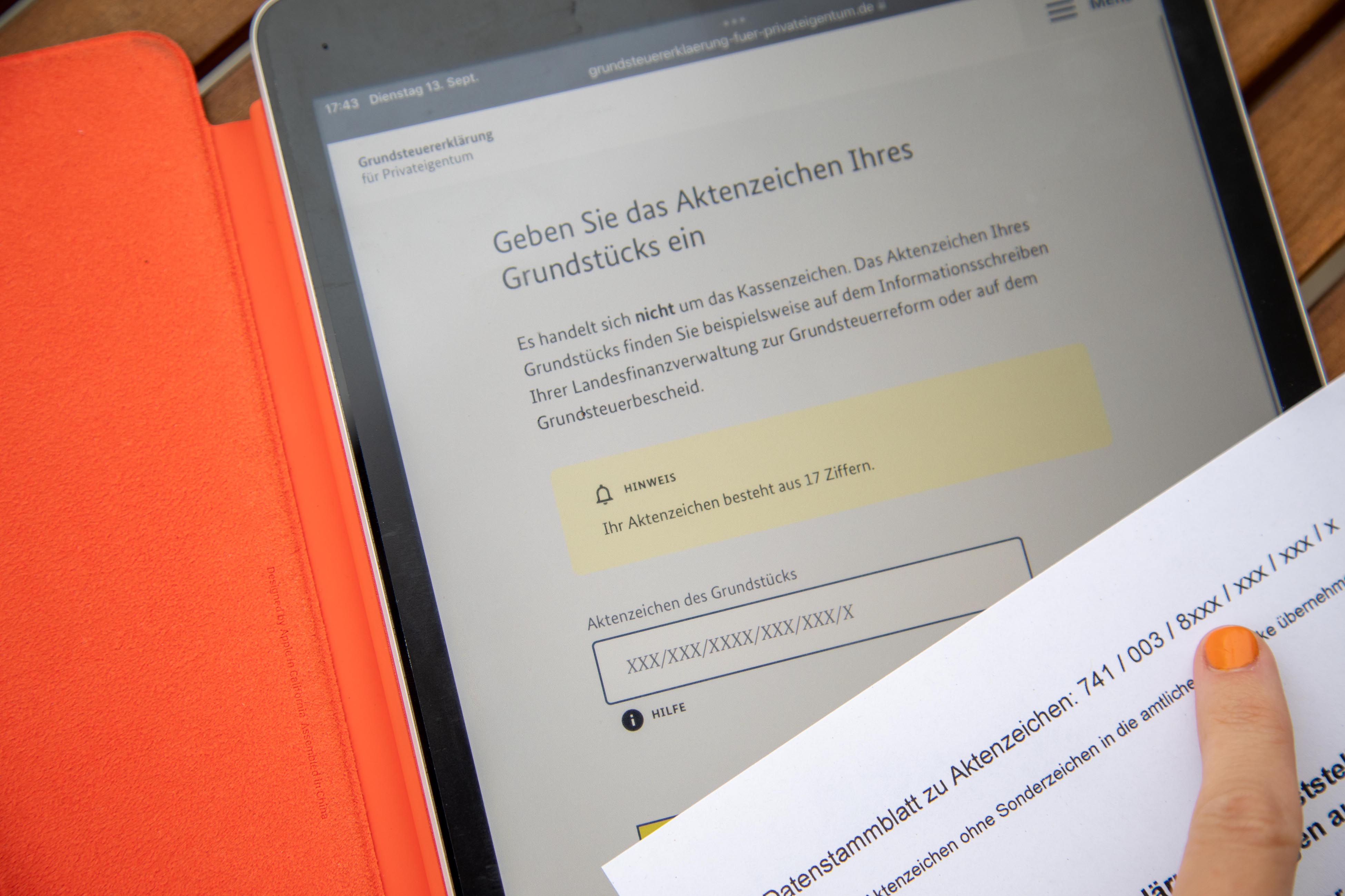 Ein Tablet liegt auf einem Holztisch; darauf geöffnet ist eine Seite des Online-Service „Grundsteuererklärung für Privateigentum“ auf der verlangt wird, dass das Aktenzeichen des Grundstücks eingegeben wird; ein Finger mit orangenem Nagellack deutet auf ein Blatt Papier auf dem das Aktenzeichen zu lesen ist