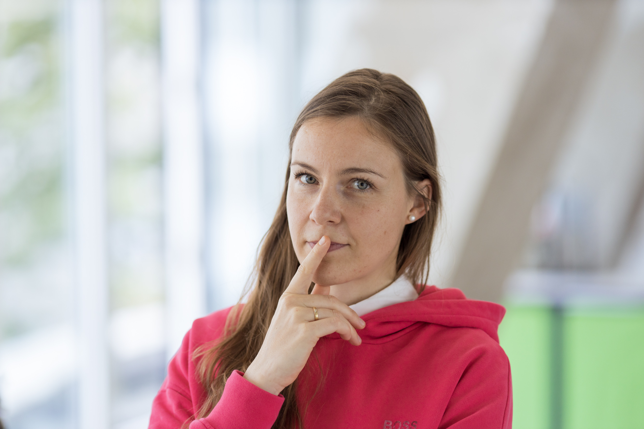 Compliance Managerin Julie in nachdenklicher Pose mit ihrem Zeigefinger über den Lippen. Sie trägt einen pinken Kapuzenpullover.