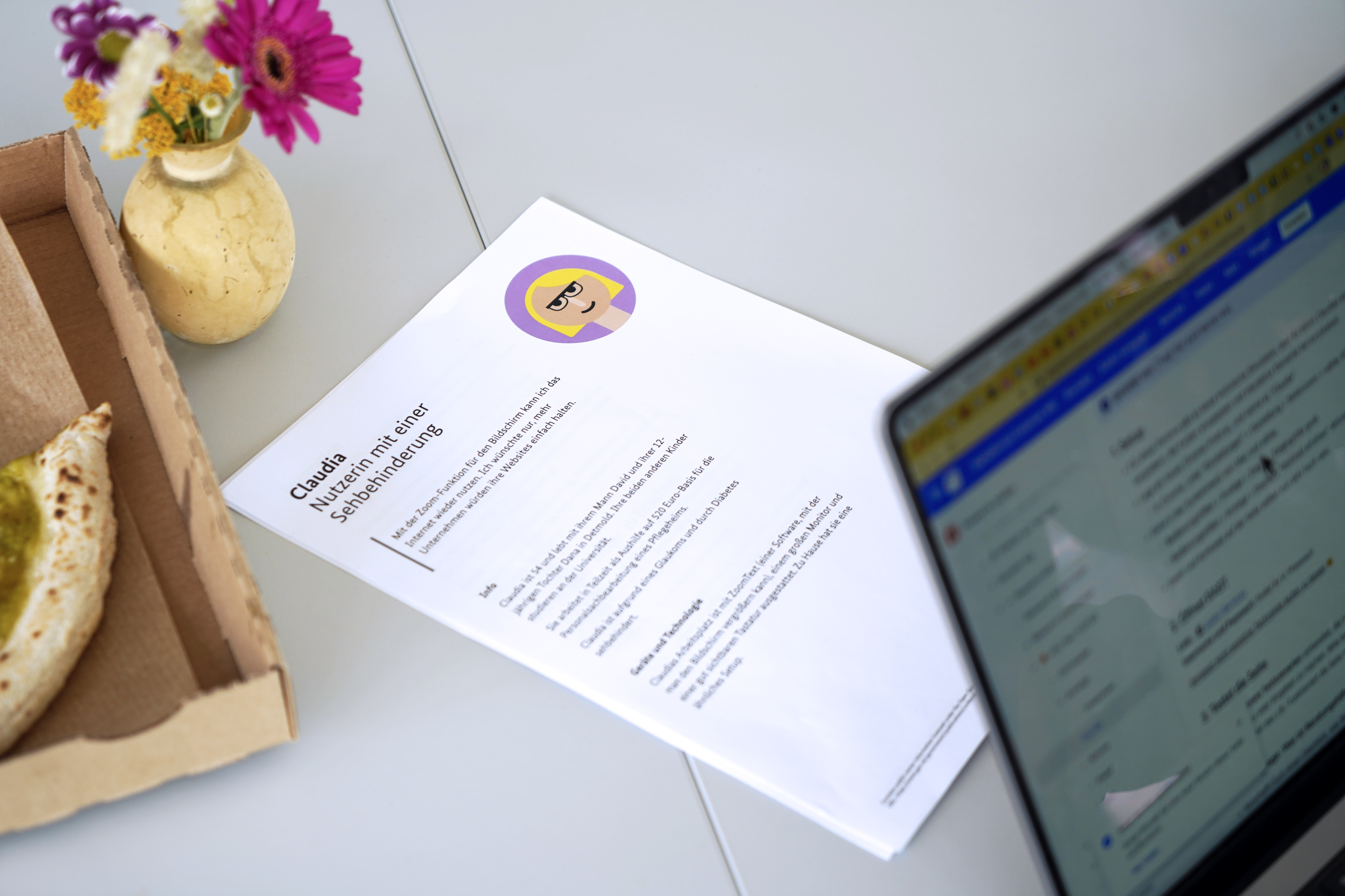 Ein Blatt mit einer Beschreibung für eine Nutzerin mit einer Sehbehinderung neben einer Pizzaschachtel und einem Laptop-Computer