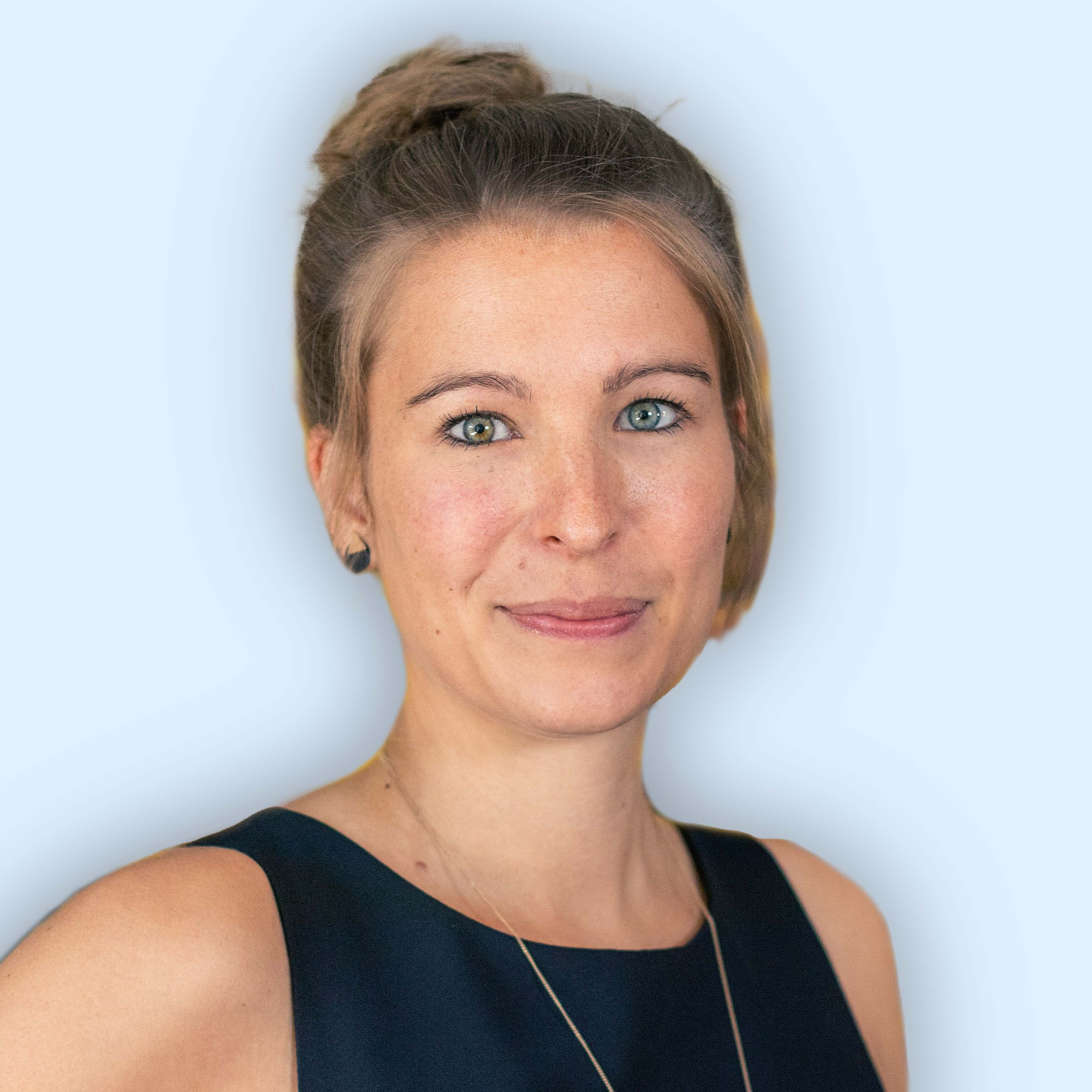 Michaela steht vor einer weißen Wand. Auf dem Portait Bild lacht Sie in die Kamera. Michaela hat eine Kette um den Hals und trägt ein Blaues Oberteil. Ihre Haare sind auf dem Kopf zu einem Dutt gebunden.