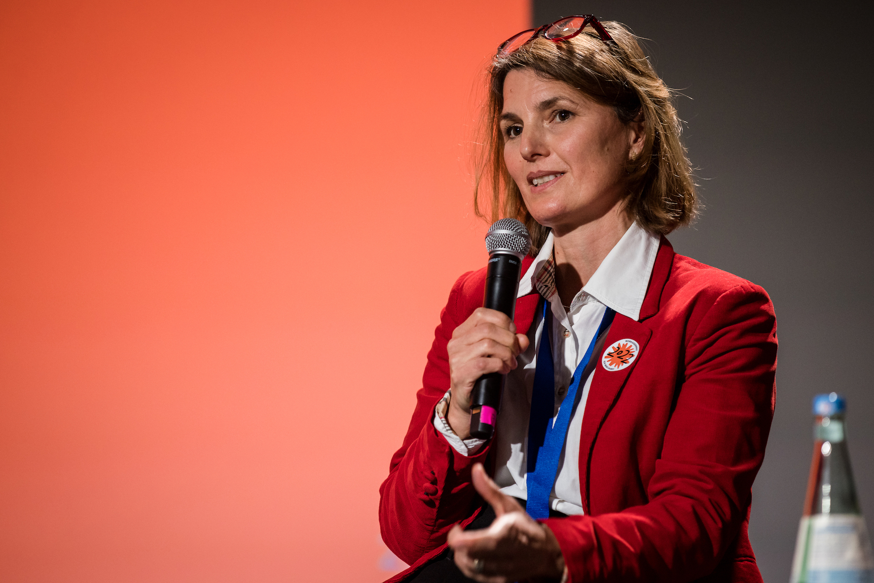 Eine weiße Frau mittleren Alters in weißer Bluse und rotem Blazer mit einem Mikrofon in der Hand und einem kleinen orange-weißen Aufkleber mit der Zahl 2022.