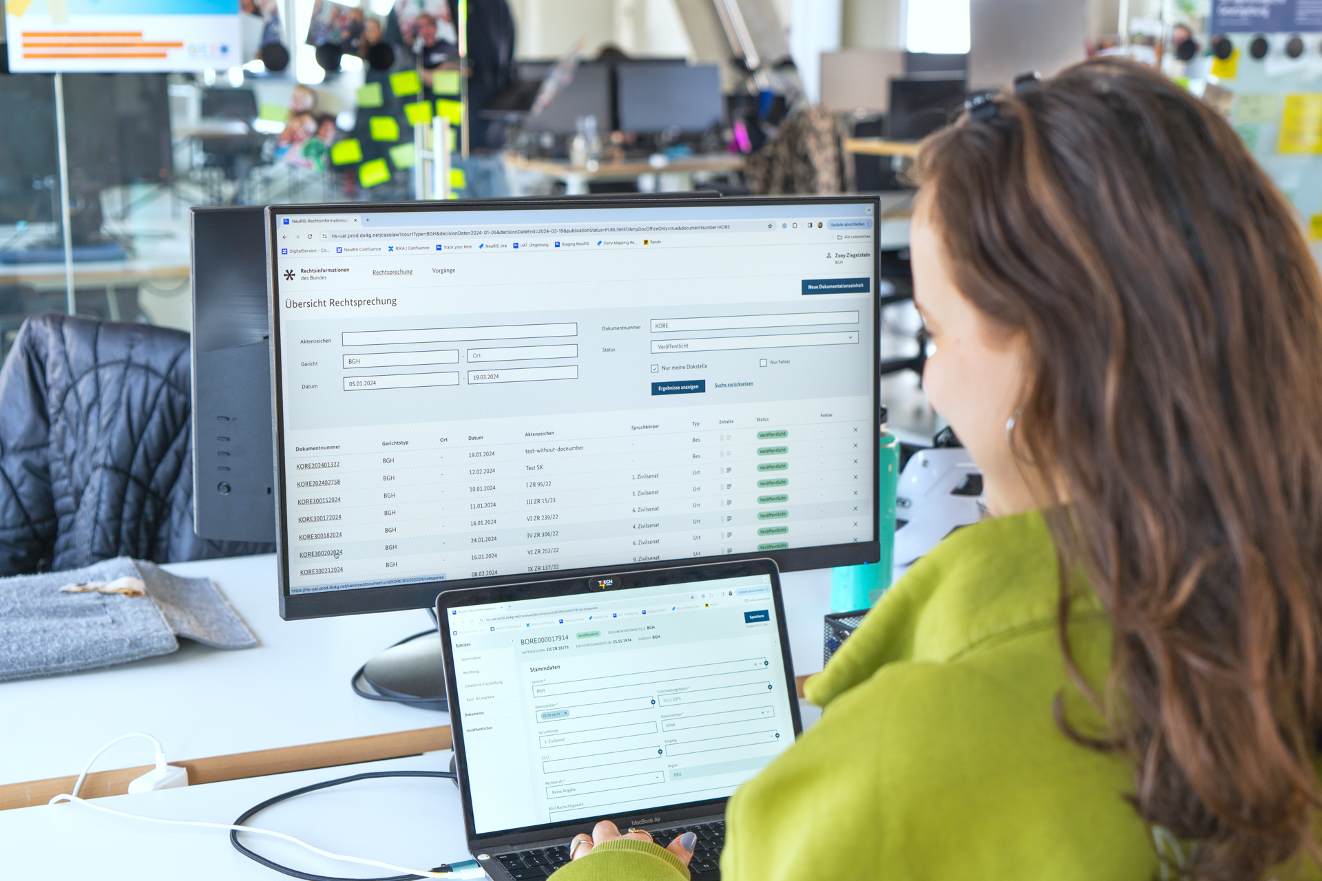 Über die Schuler einer Mitarbeiterin blickend ist auf einem großen Bildschirm sowie auf einem Laptop-Bildschirm die Nutzeroberfläche des sich im Pilotbetrieb befindenden Rechtsinformationssystems zu sehen