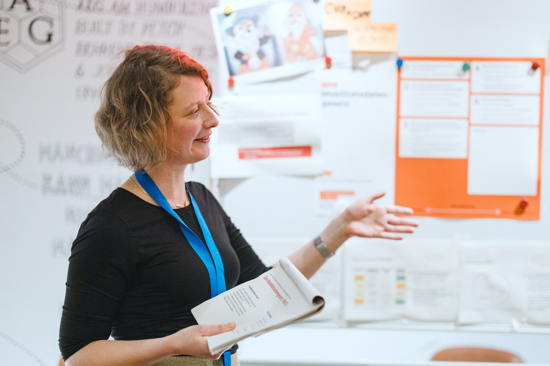 Die Autorin Audrey steht vor einer Wand, an der viele Zettel und Plakate geheftet sind; sie hält einen Vortrag zum Mobi­litäts­da­ten­gesetz