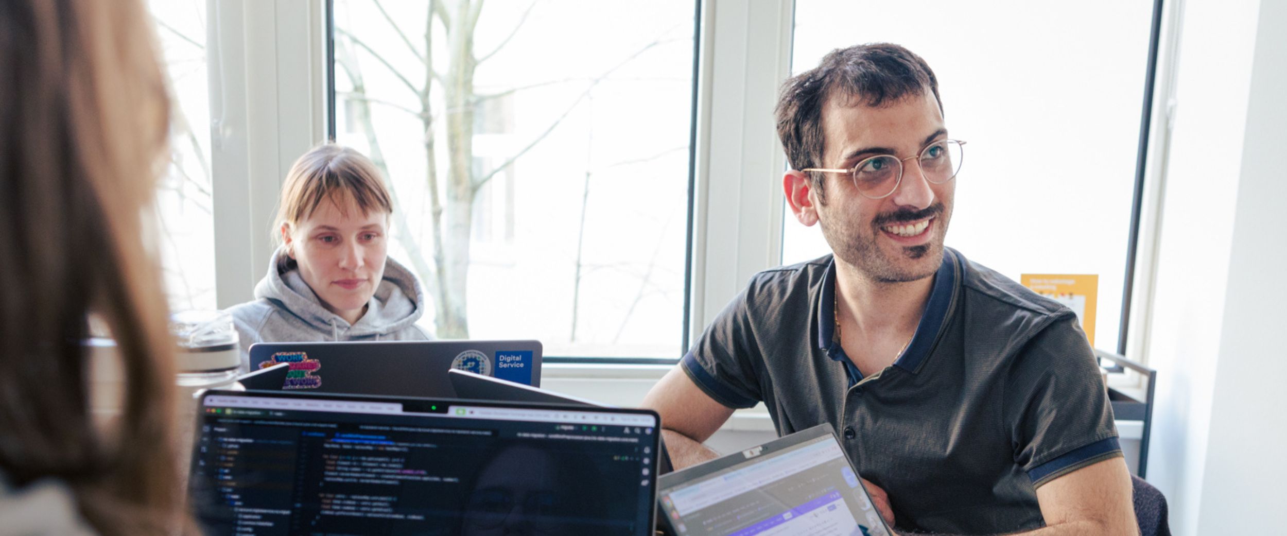 Drei Personen sitzen in einem Büro und arbeiten an ihren Laptops die vor ihnen stehen. Auf den Bildschirmen ist Code zu sehen. Im Hintergrund ist eine Fensterfront durch die Licht fällt.