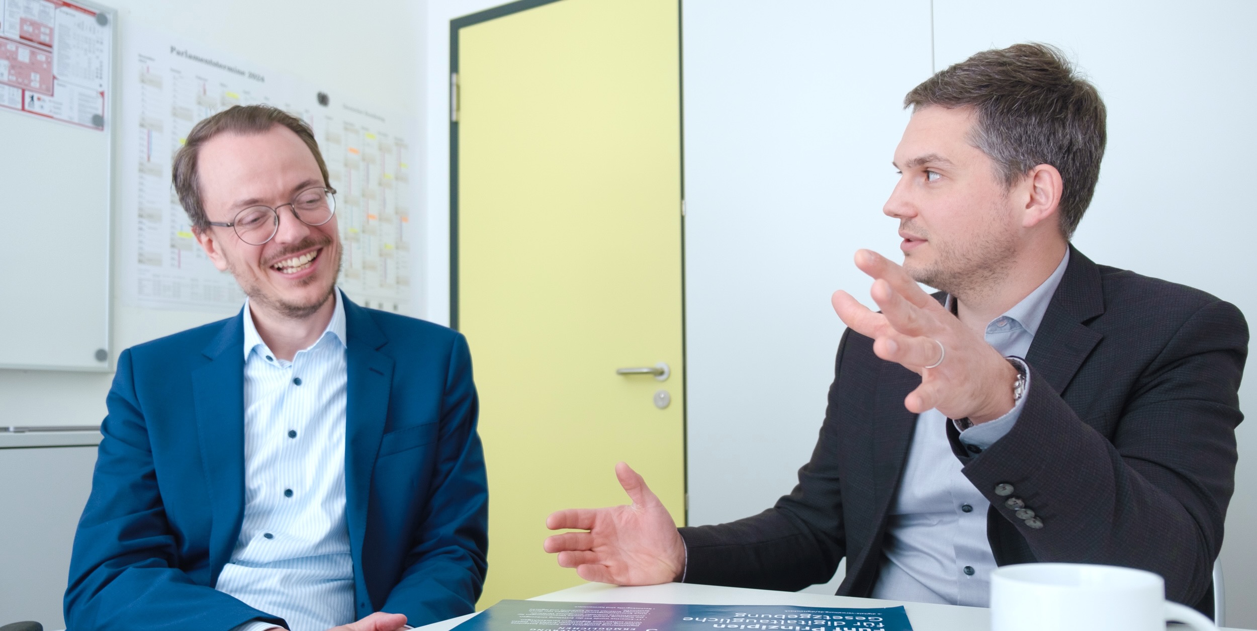 Two men in conversation, one smiling in a blue suit, the other gesticulating in a dark jacket. A door frame and pinboards in the background.