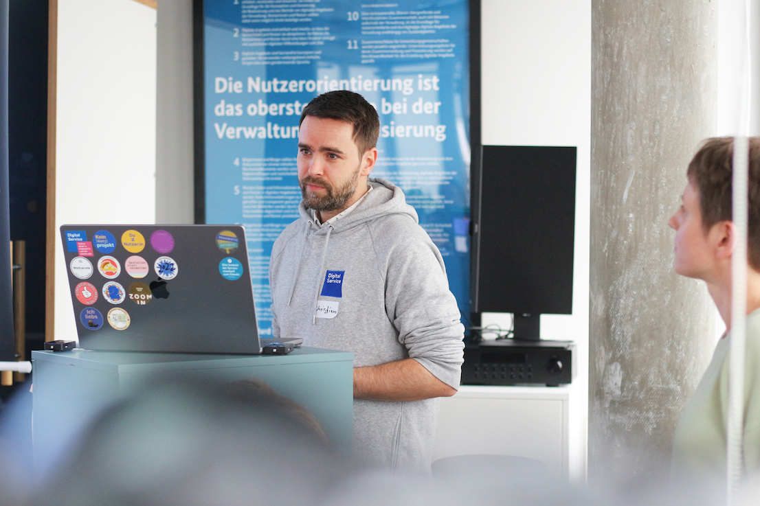 Christian Kaatz steht vor einem Podium mit einem Computer mit vielen bunten Aufklebern; im Hintergrund ist ein Plakat mit der Aufschrift „Die Nutzerzentrierung ist das oberste Prinzip bei der Verwaltungsdigitalisierung“ zu sehen