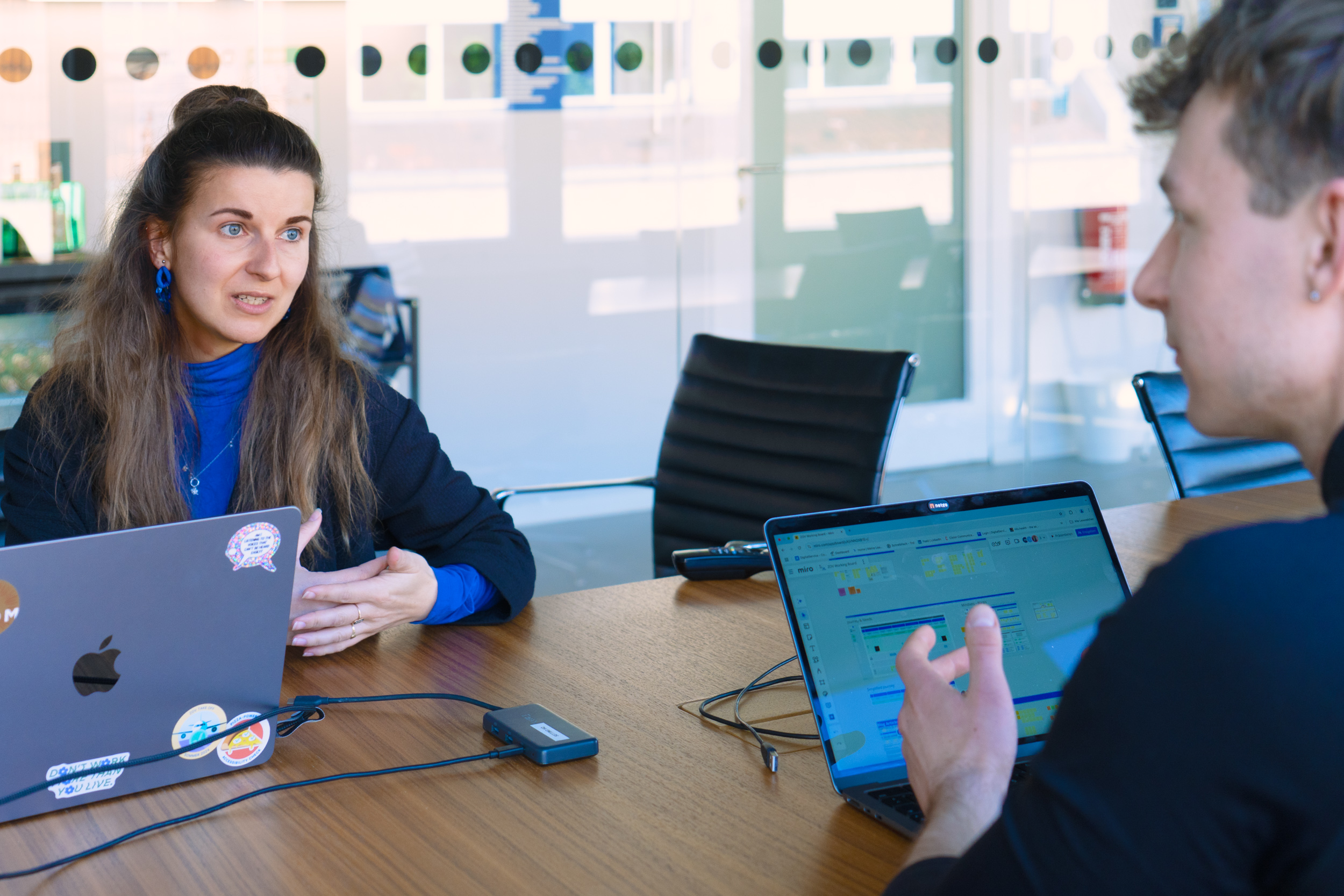 Zwei Personen sitzen sich an einem Holztisch in einem Meetingraum gegenüber und diskutieren. Beide haben geöffnete Laptops vor sich. Eine Glaswand im Hintergrund.