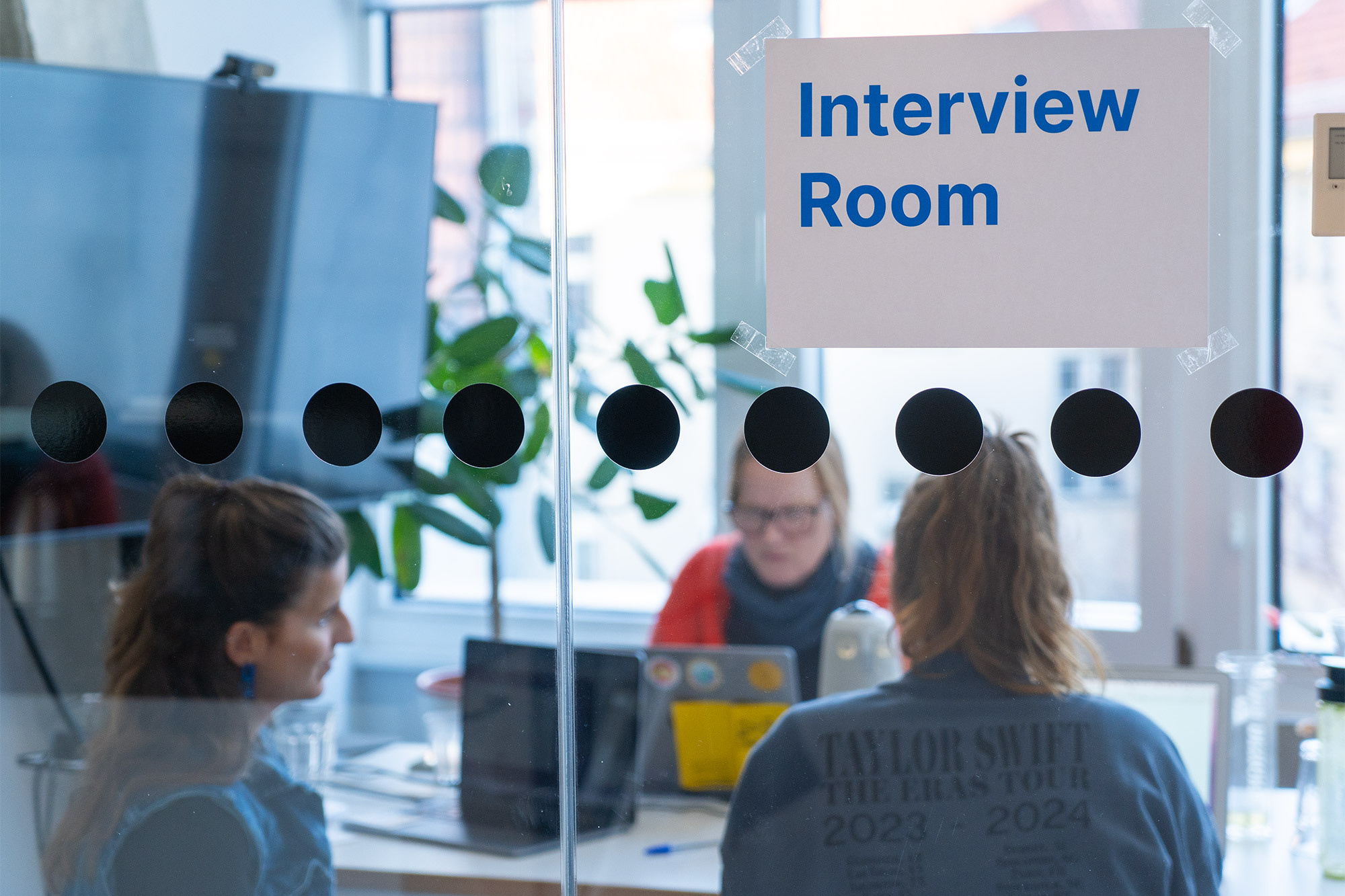 Ein Meetingraum mit Glaswand mit Schild „Interview Room“ und schwarzen Punkten auf der Scheibe. Drei Personen am Tisch mit Laptops. Im Hintergrund eine Pflanze und ein großer Bildschirm.