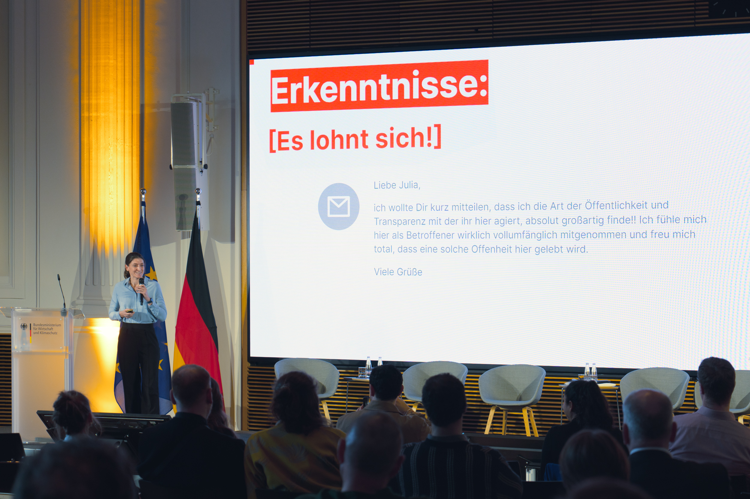 Eine Person spricht auf einer Bühne vor Publikum. Hinter ihr ist eine große Leinwand mit der Überschrift „Erkenntnisse: [Es lohnt sich!]“ und einer Nachricht mit lobenden Worten.