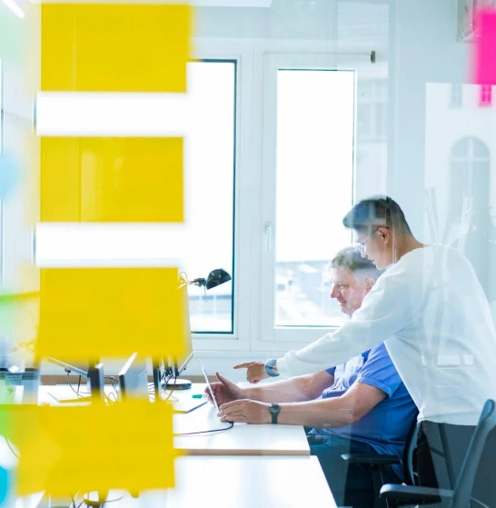 Zwei Menschen sitzen in einem Glasbüro am Computer. Auf dem Glas sind gelbe Post-It Sticker.