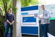 Zwei Personen stehen jeweils links und rechts neben einer Tafel und präsentieren etwas. Auf der Tafel ist ein Plakat mit der Aufschrift„Servicestandard.gov.de“, und einer Abbildung der Servicestandard Webseite mit einem QR-Code.