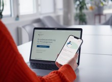 Eine Person in einem orangefarbenen Pullover hält ein Smartphone mit einer geöffneten Webseite, während im Hintergrund ein Laptop mit der Seite 