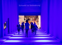 Eine Gruppe von Personen läuft in einem schräg abfallenden, tunnelartigen Zugang mit hohen Wänden nach unten in Richtung einer hell erleuchteten Garderobe; der Zugang ist in blaues Licht gehüllt; am oberen Rand des Zugangs steht „Schwelle zur Veränderung“
