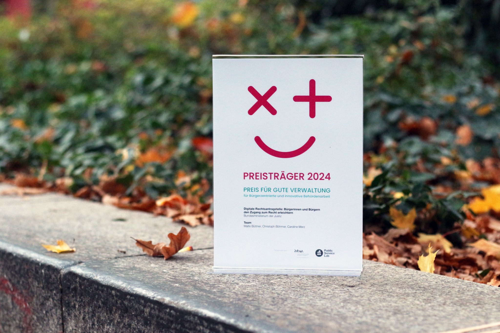 An award with the inscription „Prize Winner 2024 - Prize for Good Administration“ stands on a stone pedestal outside, surrounded by autumnal foliage.