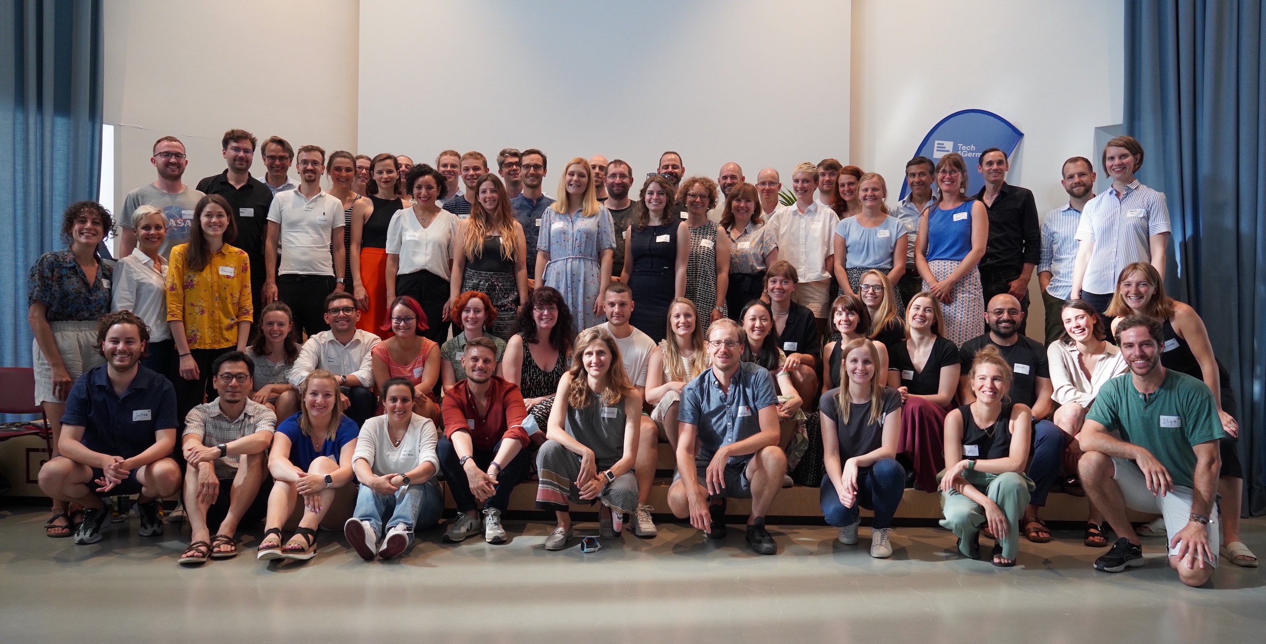 Gruppenfoto des 5. Tech4Germany Jahrgangs mit über 60 Stipendiaten und Projektpartnern. Sie stehen in einem Raum mit hohen Decken.  Links und rechts gibt hängen Vorhänge.