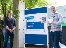 Zwei Personen stehen jeweils links und rechts neben einer Tafel und präsentieren etwas. Auf der Tafel ist ein Plakat mit der Aufschrift„Servicestandard.gov.de“, und einer Abbildung der Servicestandard Webseite mit einem QR-Code.