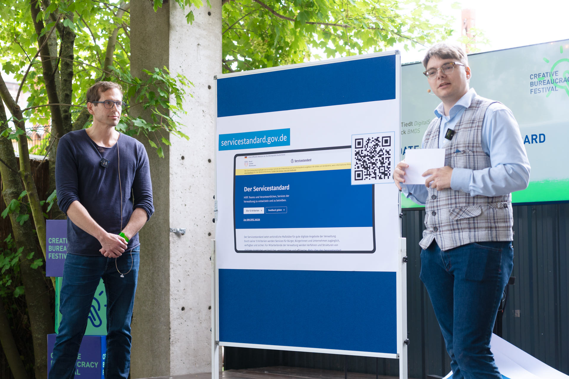 Zwei Personen stehen jeweils links und rechts neben einer Tafel und präsentieren etwas. Auf der Tafel ist ein Plakat mit der Aufschrift„Servicestandard.gov.de“, und einer Abbildung der Servicestandard Webseite mit einem QR-Code.
