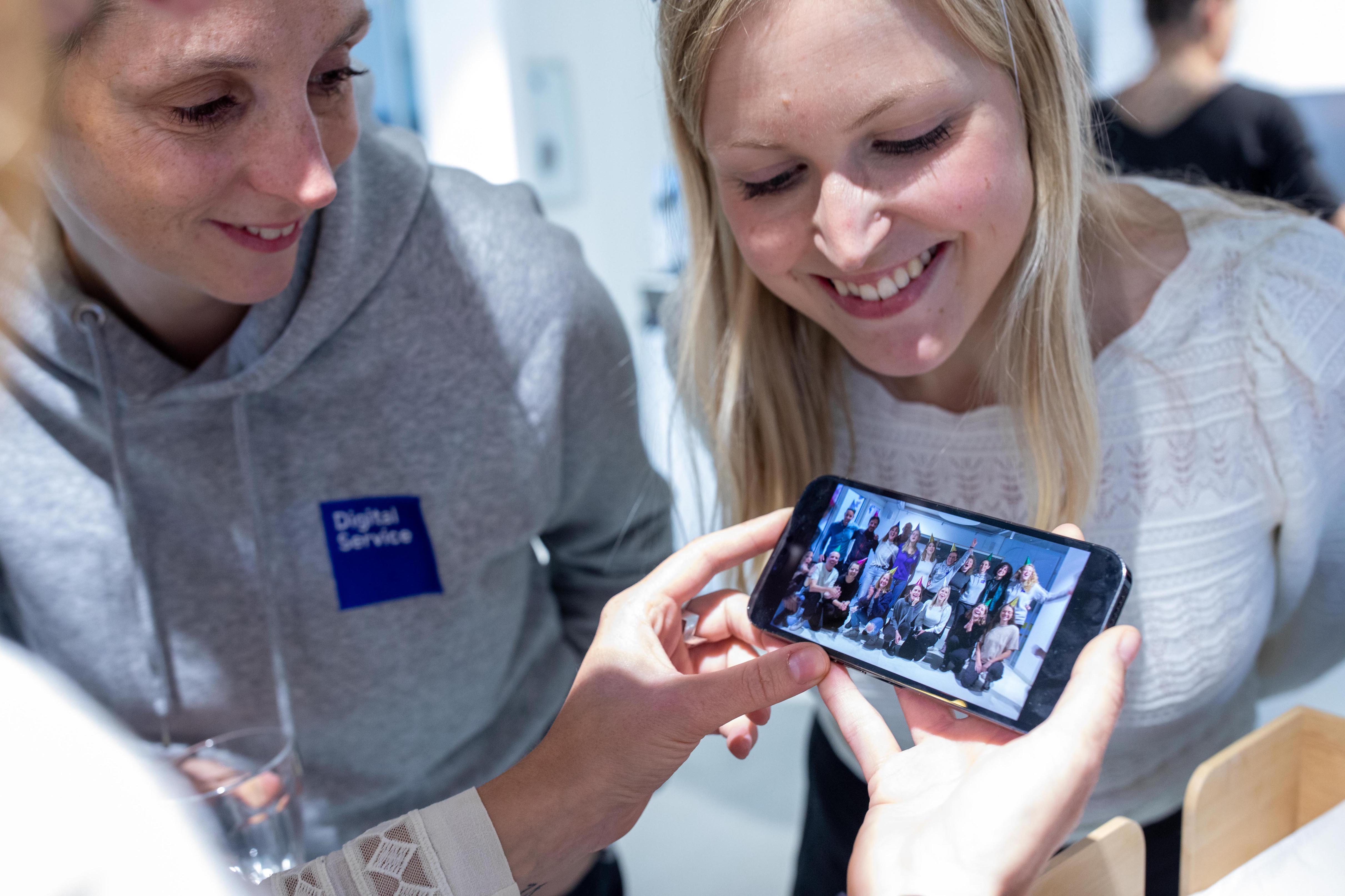 Two smiling people are looking at a smartphone showing a group photo. One of the people is wearing a gray hoodie with the “Digital Service” logo.