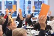 Eine Gruppe von Menschen in einem Sitzungssaal hebt orangefarbene Karten bei der Verabschiedung der DIN SPEC 66336. Die Personen sitzen an Tischen mit Getränken und Dokumenten, während sie aktiv an der Versammlung teilnehmen.