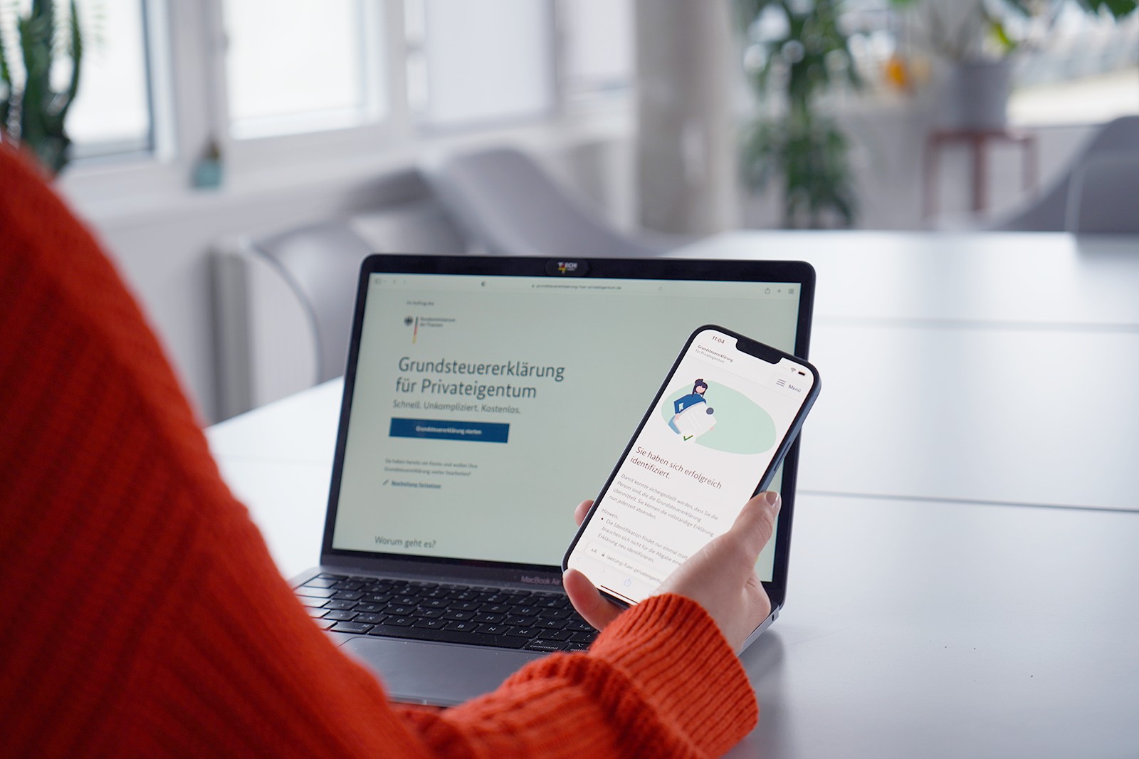 A person in an orange sweater holds a smartphone with a website open, while a laptop with the page “Property tax declaration for private property” can be seen on the screen in the background.