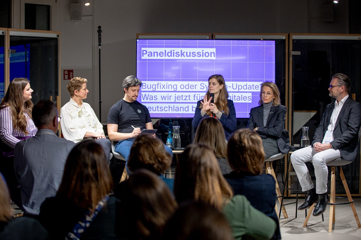 A panel discussion with five participants on a stage. Christina Lang speaks into the microphone while the audience listens. In the background, a screen shows the title of the discussion “Bug fixing or system update? What we need now for a digital Germany”