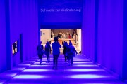 Eine Gruppe von Personen läuft in einem schräg abfallenden, tunnelartigen Zugang mit hohen Wänden nach unten in Richtung einer hell erleuchteten Garderobe; der Zugang ist in blaues Licht gehüllt; am oberen Rand des Zugangs steht „Schwelle zur Veränderung“