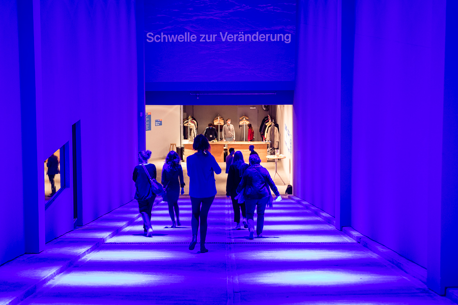 A group of people walk down a sloping, tunnel-like entrance with high walls towards a brightly lit checkroom; the entrance is bathed in blue light; “Threshold of Change” is written at the top of the entrance