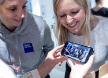 Zwei lachende Personen schauen auf ein Smartphone, das ein Gruppenfoto zeigt. Eine der Personen trägt einen grauen Hoodie mit dem Logo „DigitalService“.