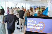 Im Vordergrund ist auf einem Laptopbildschirm zu lesen, dass mehr als 150 Mitarbeitende nun beim DigitalService arbeiten; im Hintergrund sind unscharf zahlreiche Mitarbeitende zu erkennen, die gemeinsam feiern