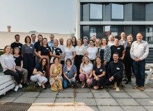 Eine Gruppe von Menschen, der 5. Jubiläumsjahrgang des Work4Germany Fellowships steht auf einer Terrasse aus Steinquadern. Links und rechts sind Bänke, im Hintergrund sind Bürogebäude zu sehen.