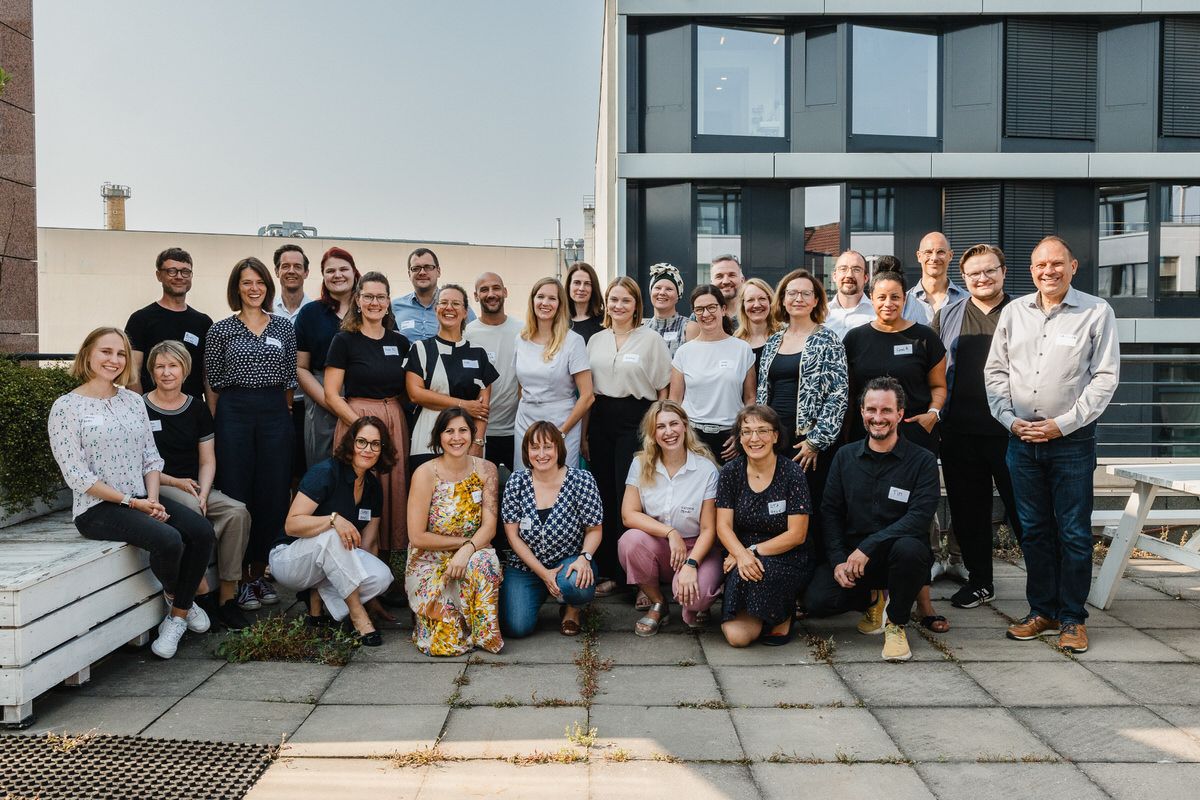 A group of people, the 5th anniversary class of the Work4Germany Fellowship, stands on a terrace made of stone blocks. There are benches to the left and right, and office buildings can be seen in the background.