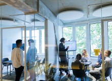 Personen im Meeting im ersten eigenen Büro im August 2021. Das Büro ist mit einer Glaswand abgetrennt. Im Büro präsentiert eine Person auf einem Bildschirm, drei andere sitzen und hören zu.