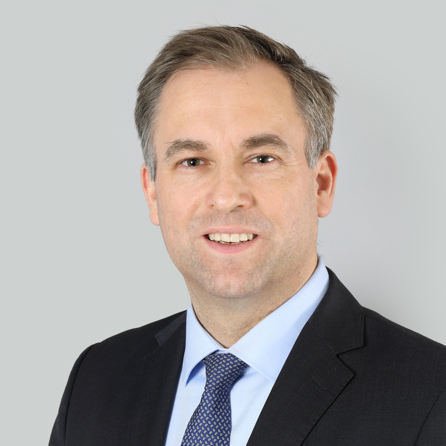 Supervisory Board member Thomas Rieks stands in front of a light gray wall and looks into the camera with a slight smile; he is wearing a black jacket, a light blue shirt and a dark blue patterned tie; he has short brown-grey mottled hair