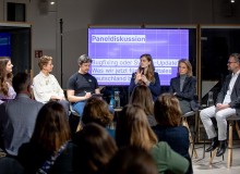 Eine Paneldiskussion mit fünf Teilnehmenden auf einer Bühne. Christina Lang spricht ins Mikrofon, während das Publikum zuhört. Im Hintergrund zeigt eine Leinwand den Titel der Diskussion „Bugfixing oder System-Update? Was wir jetzt für ein digitales Deutschland brauchen“