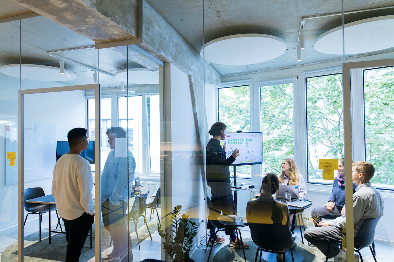 People in a meeting in their first own office in August 2021. The office is partitioned off with a glass wall. In the office, one person presents on a screen, three others sit and listen.