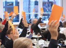Eine Gruppe von Menschen in einem Sitzungssaal hebt orangefarbene Karten bei der Verabschiedung der DIN SPEC 66336. Die Personen sitzen an Tischen mit Getränken und Dokumenten, während sie aktiv an der Versammlung teilnehmen.