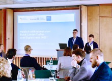 Zwei Personen stehen in einem Raum und präsentieren etwas, auf einer Leinwand hinter ihnen steht: „Herzlich willkommen zum Bund-Länder-Treffen zum Digitalcheck 2025!“. Im Vordergrund sitzen weitere Personen und hören zu.